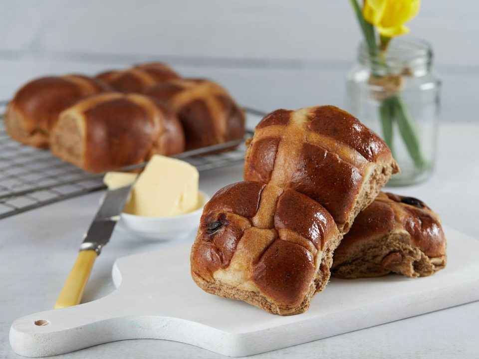 Three delicious hot cross buns in the foreground along with a butter knife and butter, and four hot cross buns on a backing tray in the background.