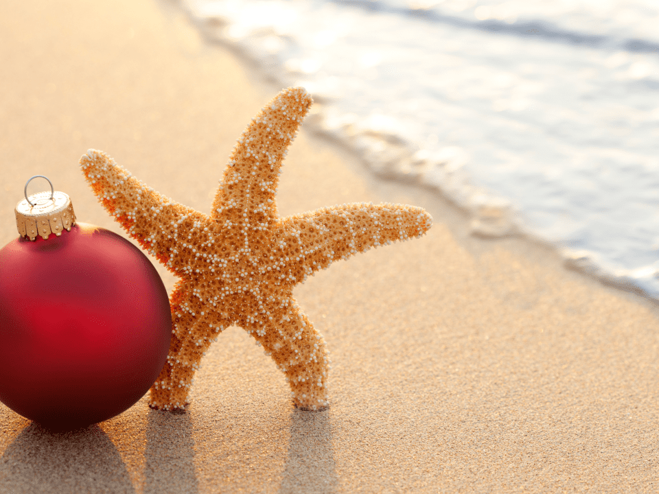 On the beach with the sea foam in the background, stands a gold colored star fish next to a red Christmas tree ball ornament.